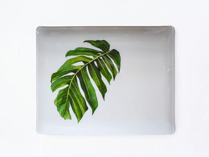 A rectangular white acrylic serving tray with rounded corners, featuring a printed green monstera leaf design in the center, set against a plain white background.