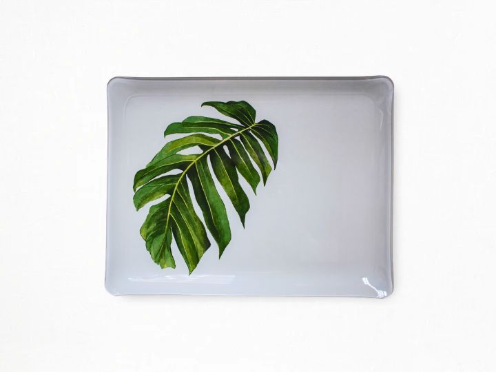 A rectangular white acrylic serving tray with rounded corners, featuring a printed green monstera leaf design on the left side, shown against a plain white background.